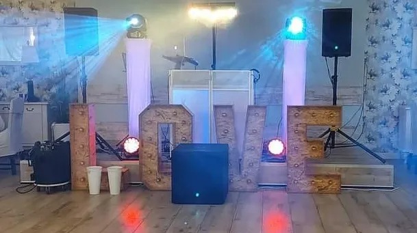 Dekoracja sali z podświetlanymi literami LOVE, sprzętem nagłośnieniowym i oświetleniem scenicznym, w tle konsoleta DJ-a