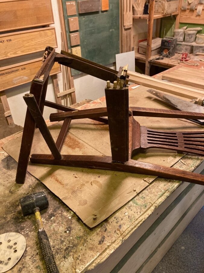 Rozłożone krzesło z ciemnego drewna na warsztatowym stole, widoczne elementy konstrukcyjne, młotek i papier ścierny, ślady napraw i renowacji.