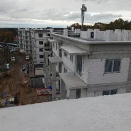 Budynek wielorodzinny w trakcie budowy, widoczne surowe elewacje z bloczków, okna zabezpieczone folią, plac budowy z maszynami i materiałami, pochmurne niebo.