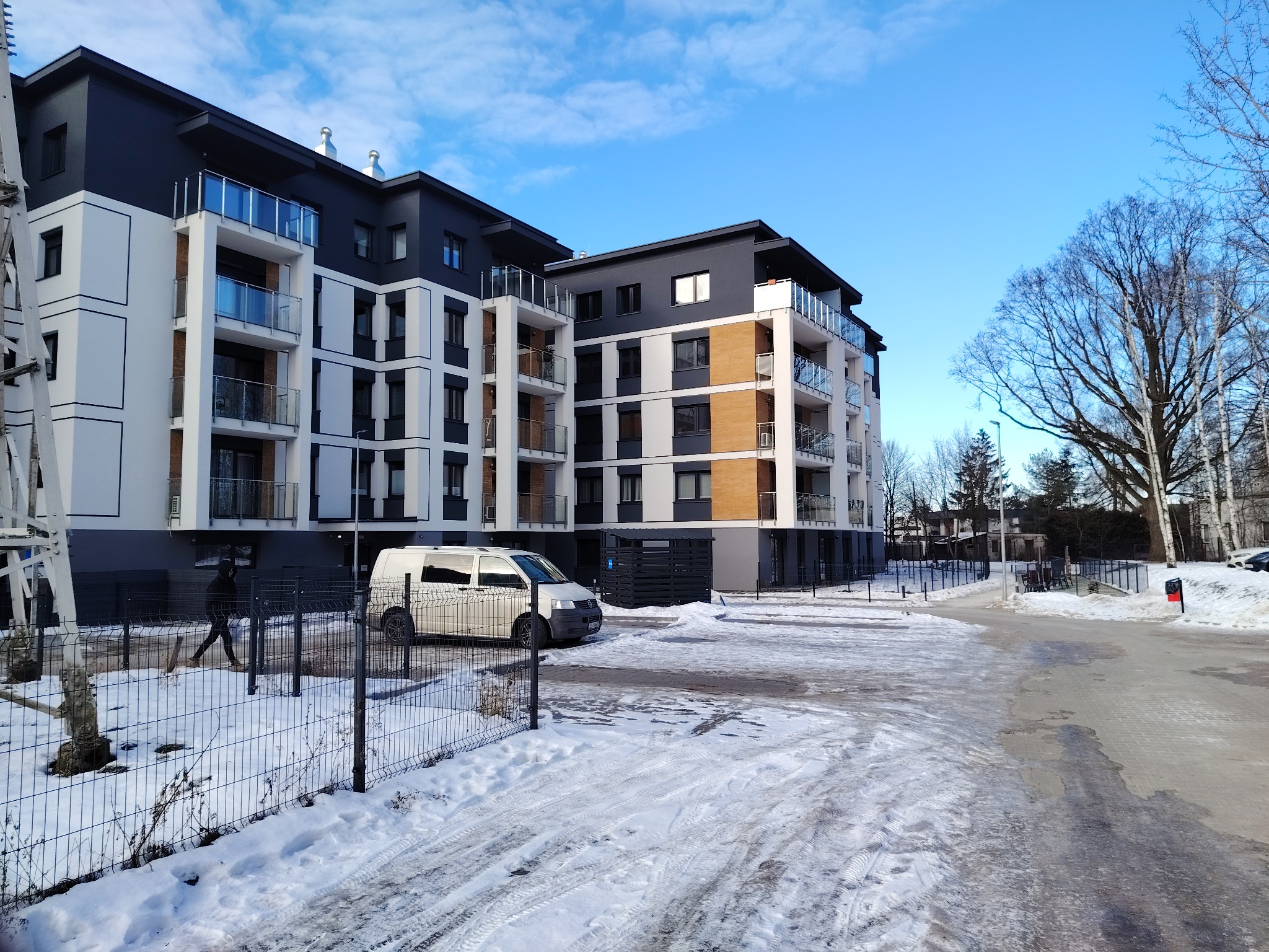 Nowoczesny budynek wielorodzinny z balkonami i elewacją w odcieniach bieli, szarości i czerni, widziany w zimowej scenerii z zaparkowanym busem.