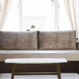 Dwukolorowa sofa z wzorzystymi poduszkami i małym stolikiem kawowym na tle okna z kotem, w jasnym, minimalistycznym wnętrzu.