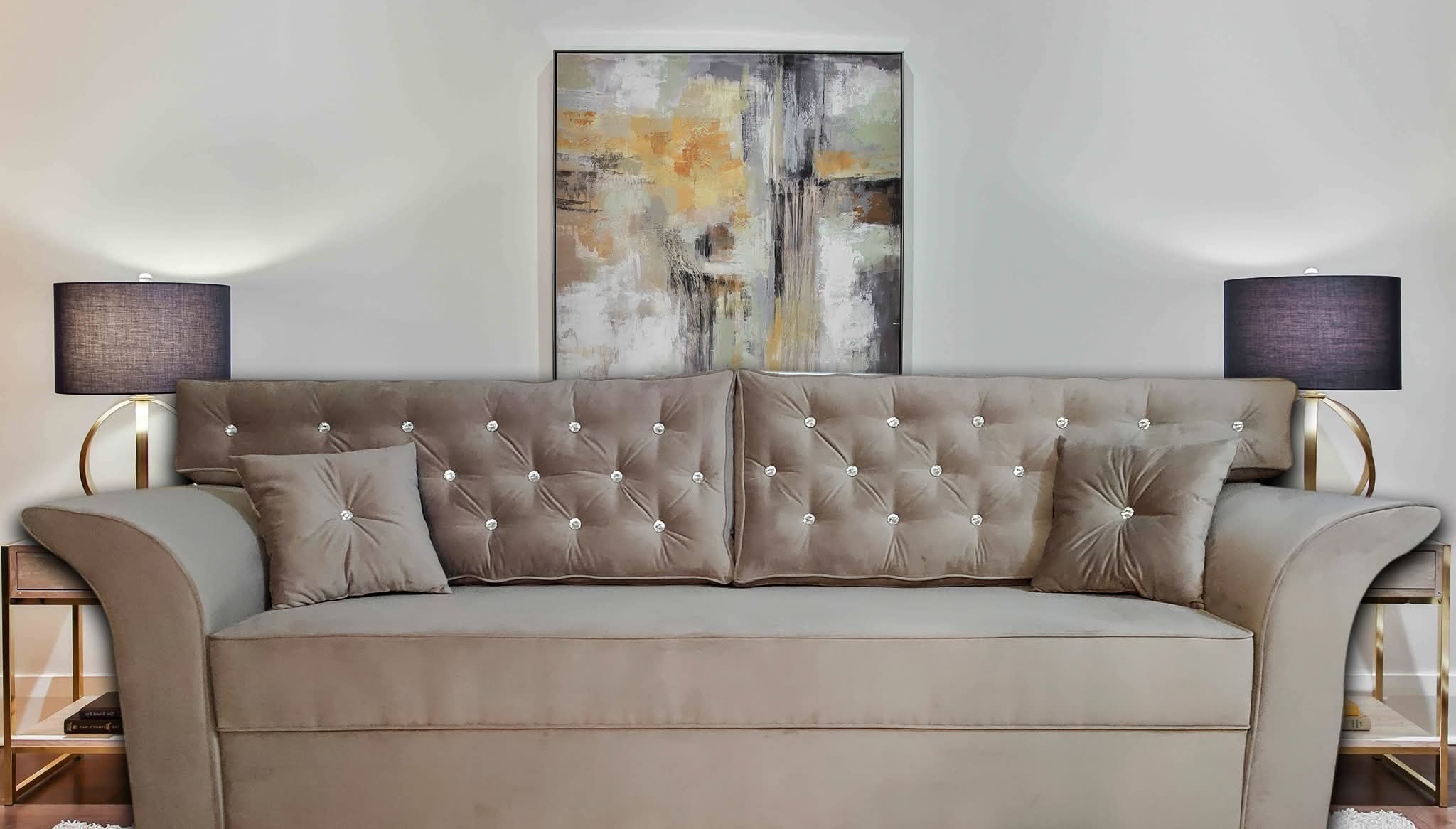 Beżowa sofa tapicerowana z guzikami, dekoracyjne poduszki, lampki nocne i abstrakcyjny obraz na ścianie tworzą elegancką aranżację wnętrza.