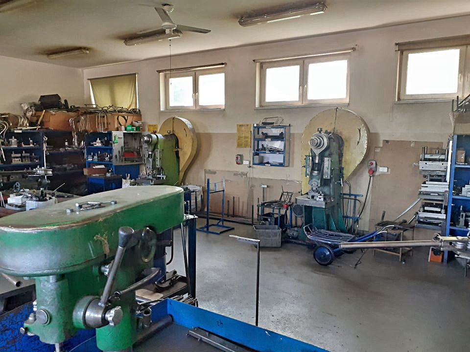 Warsztat ślusarski z zielonymi maszynami do obróbki metali, widoczne regały z narzędziami i materiałami, surowe oświetlenie.