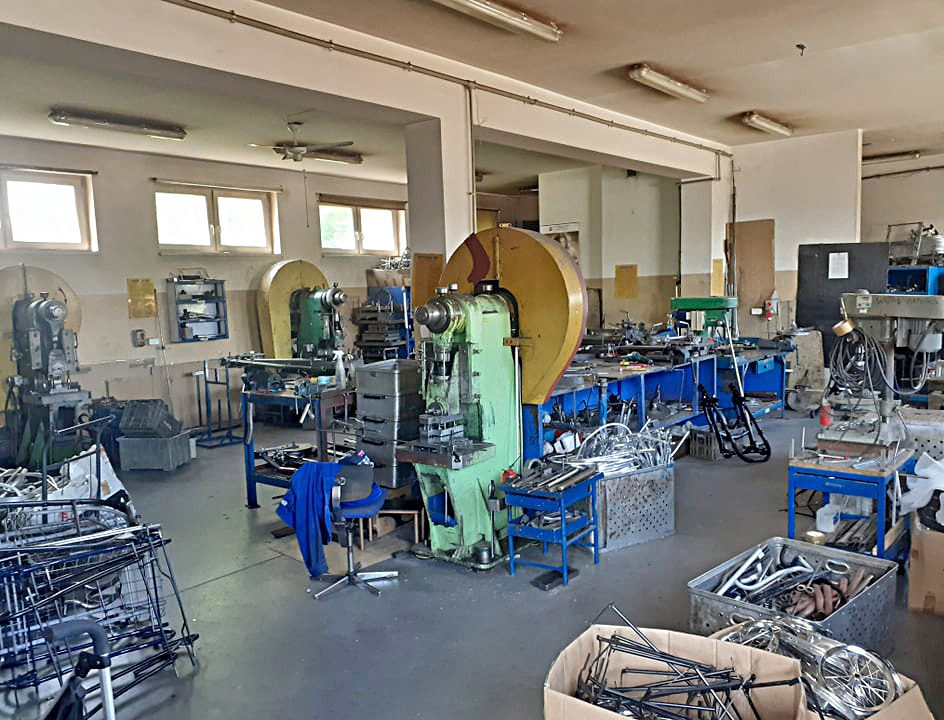 Wnętrze warsztatu z maszynami do obróbki metali, widoczne liczne narzędzia i półprodukty metalowe, w tym elementy konstrukcyjne i ramy rowerowe.