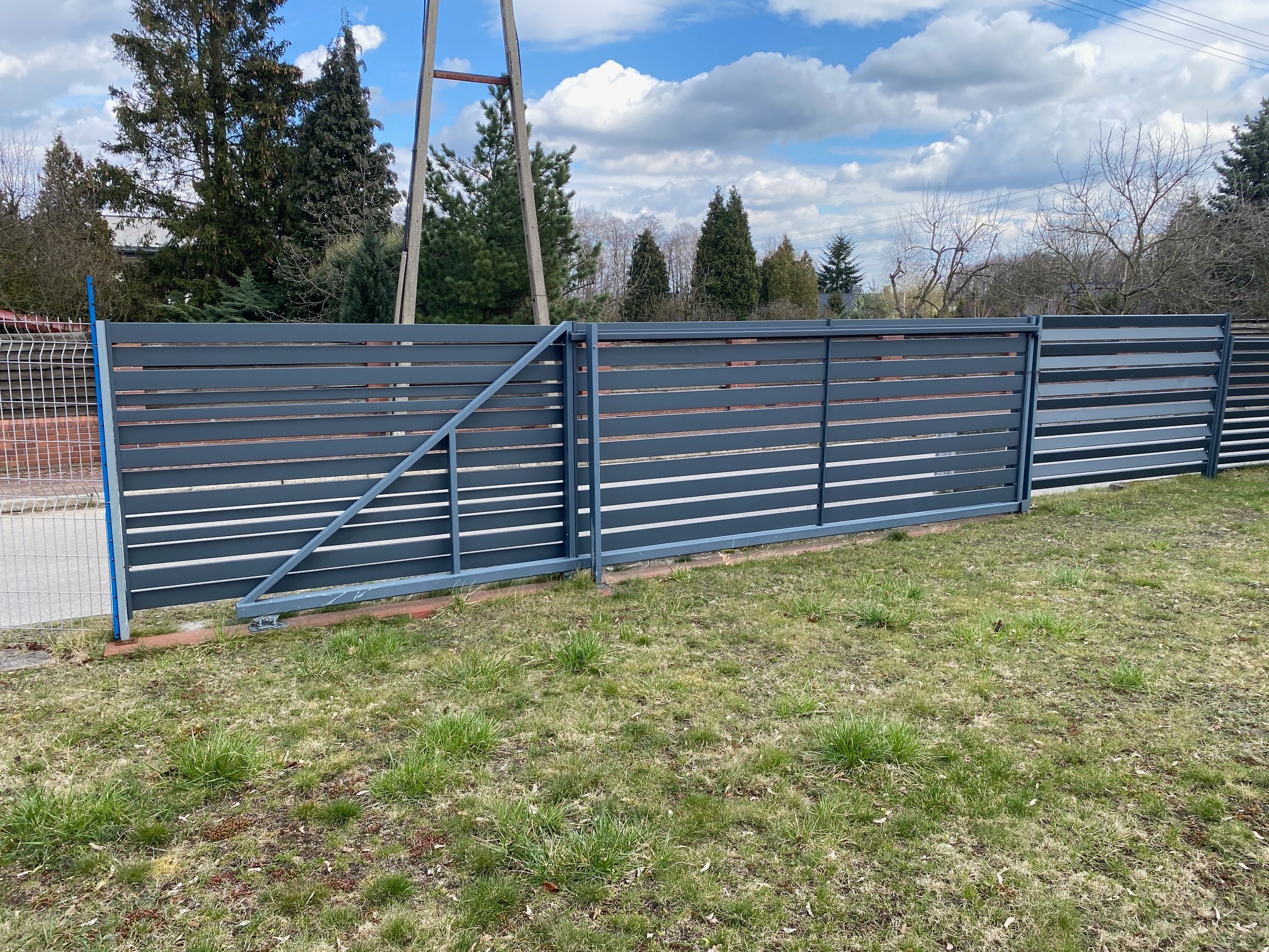 Nowoczesne ogrodzenie metalowe z poziomych paneli w kolorze grafitowym, obejmujące bramę przesuwną z widocznym mechanizmem jezdnym, na tle trawnika i drzew.