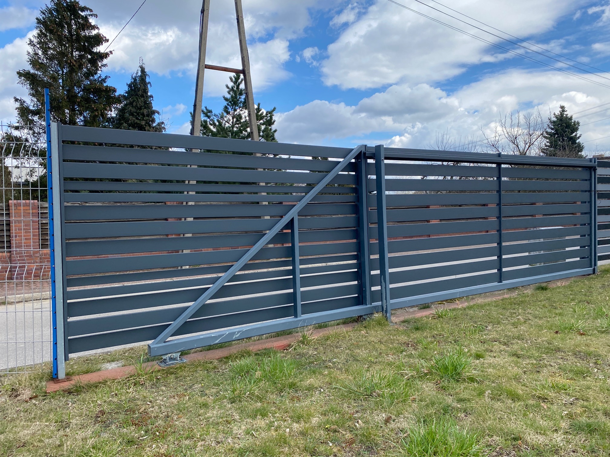 Nowoczesna, przesuwna brama wjazdowa z poziomych, szarych profili, widoczna z zewnątrz posesji, z fragmentem ogrodzenia z siatki po lewej stronie i słupem elektrycznym w tle.