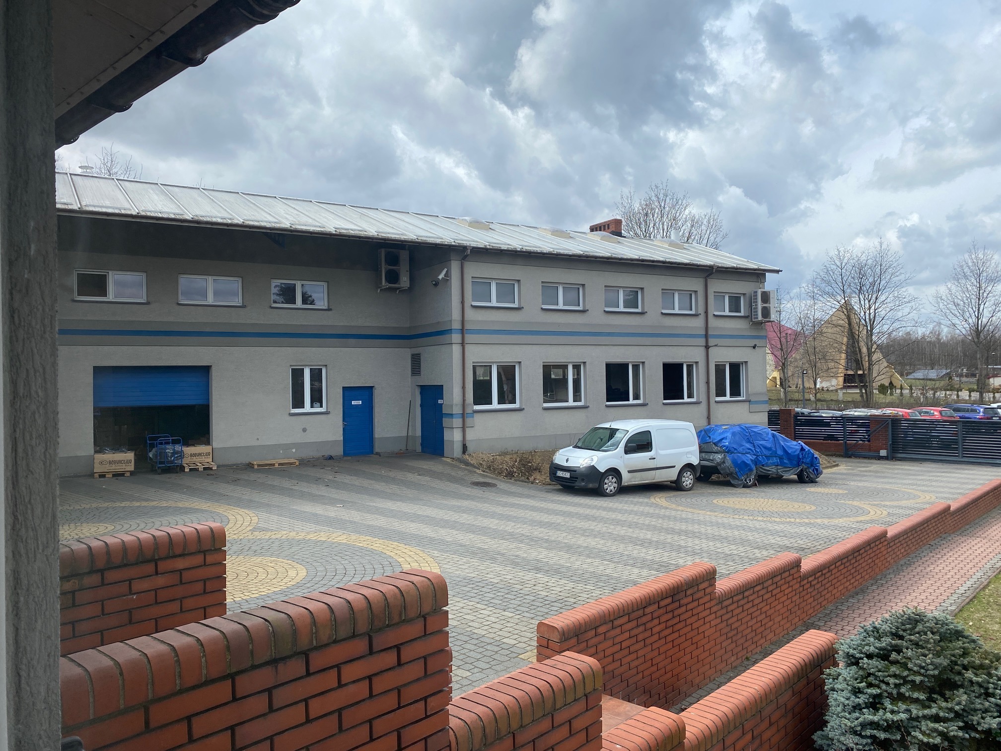 Dwukondygnacyjny budynek z szarą elewacją i niebieskimi akcentami, widoczny zza ceglanego murku, z zaparkowanym białym vanem i pojazdem przykrytym plandeką na wybrukowanym placu.
