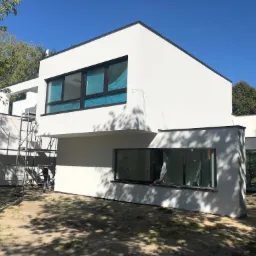 Nowoczesny, biały dom w trakcie wykończenia elewacji, z widocznymi rusztowaniami i folią ochronną na oknach, otoczony drzewami i piaszczystym terenem.