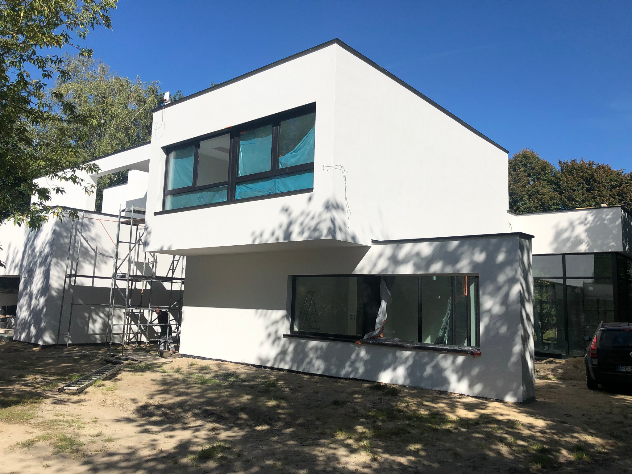 Nowoczesny, biały dom w trakcie wykończenia elewacji, z widocznymi rusztowaniami i folią ochronną na oknach, otoczony drzewami i piaszczystym terenem.