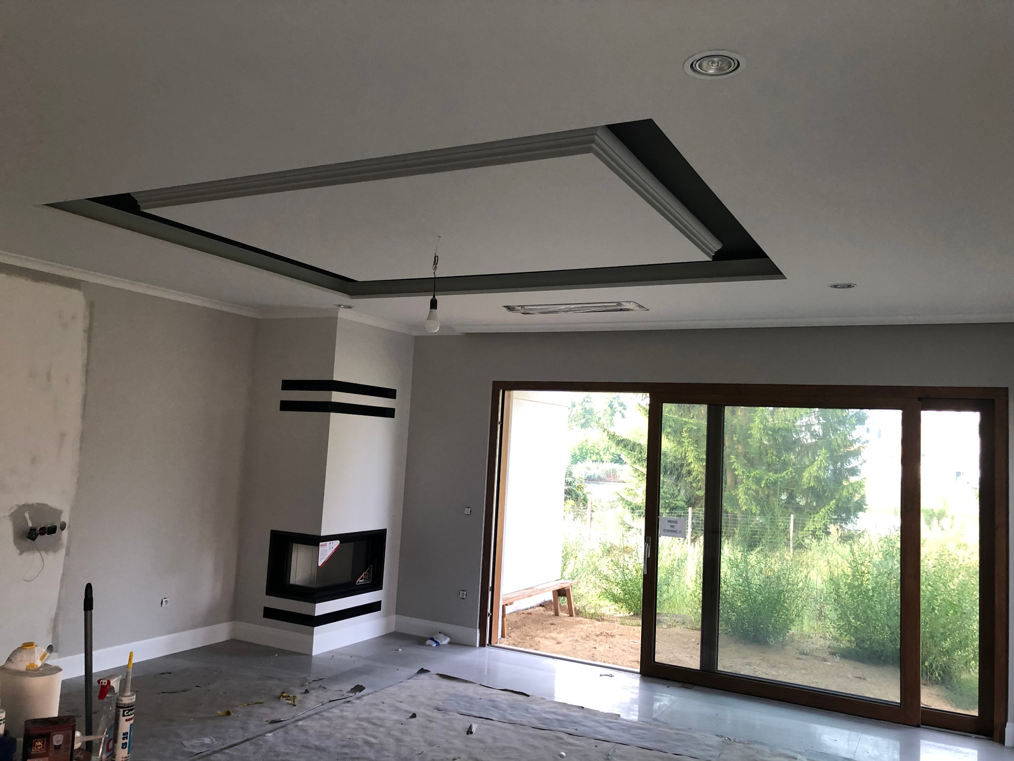 Wnętrze pokoju z kominkiem w trakcie prac wykończeniowych: pomalowane ściany na szaro, sufit z ozdobnym podwieszanym elementem, kominek z czarnymi pasami, przesuwne drzwi balkonowe z widokiem...