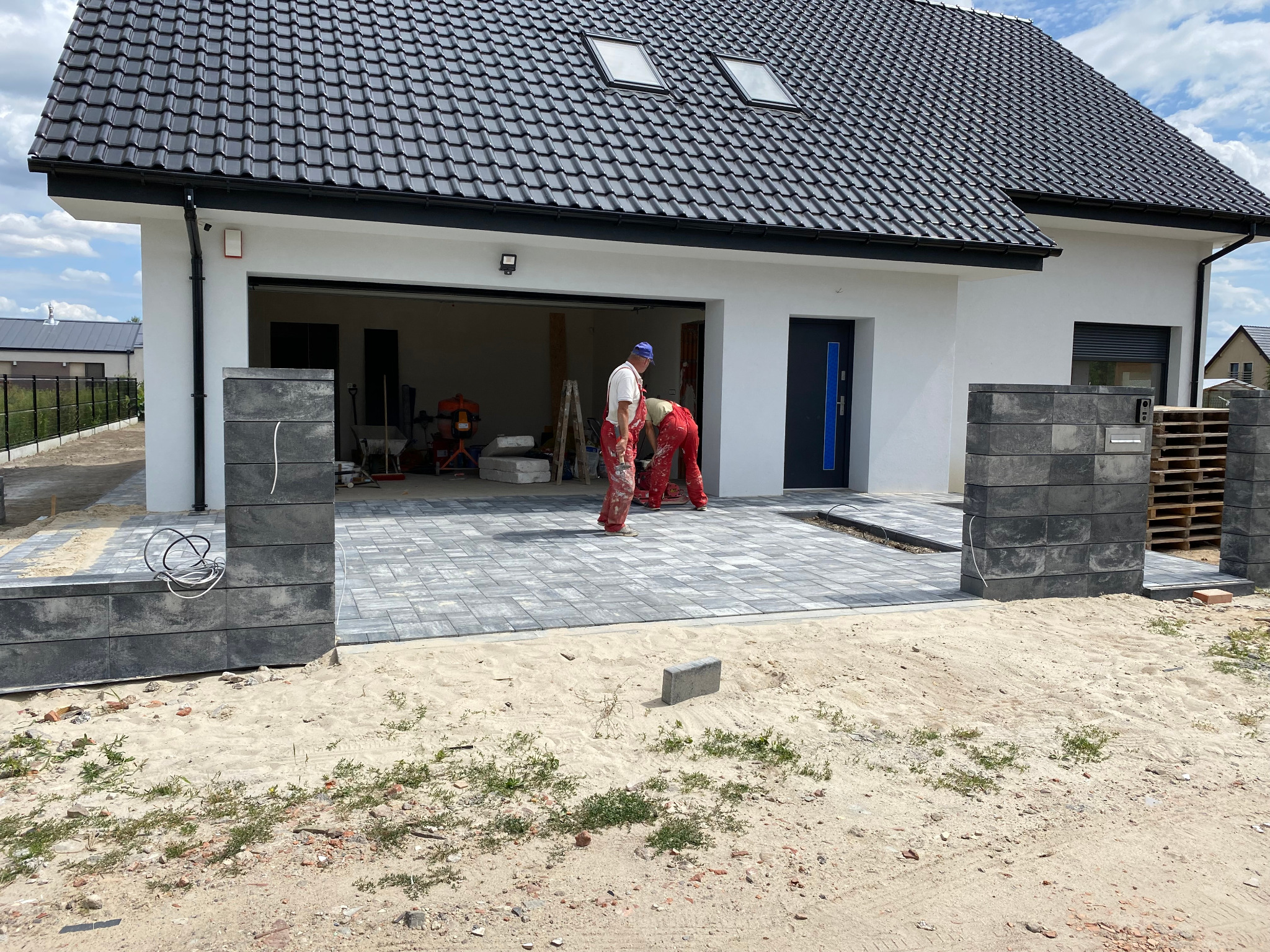 Dwóch pracowników w czerwonych kombinezonach układa szarą kostkę brukową przed nowoczesnym, białym domem z czarnym dachem, widoczne elementy piasku i niedokończonego krajobrazu.