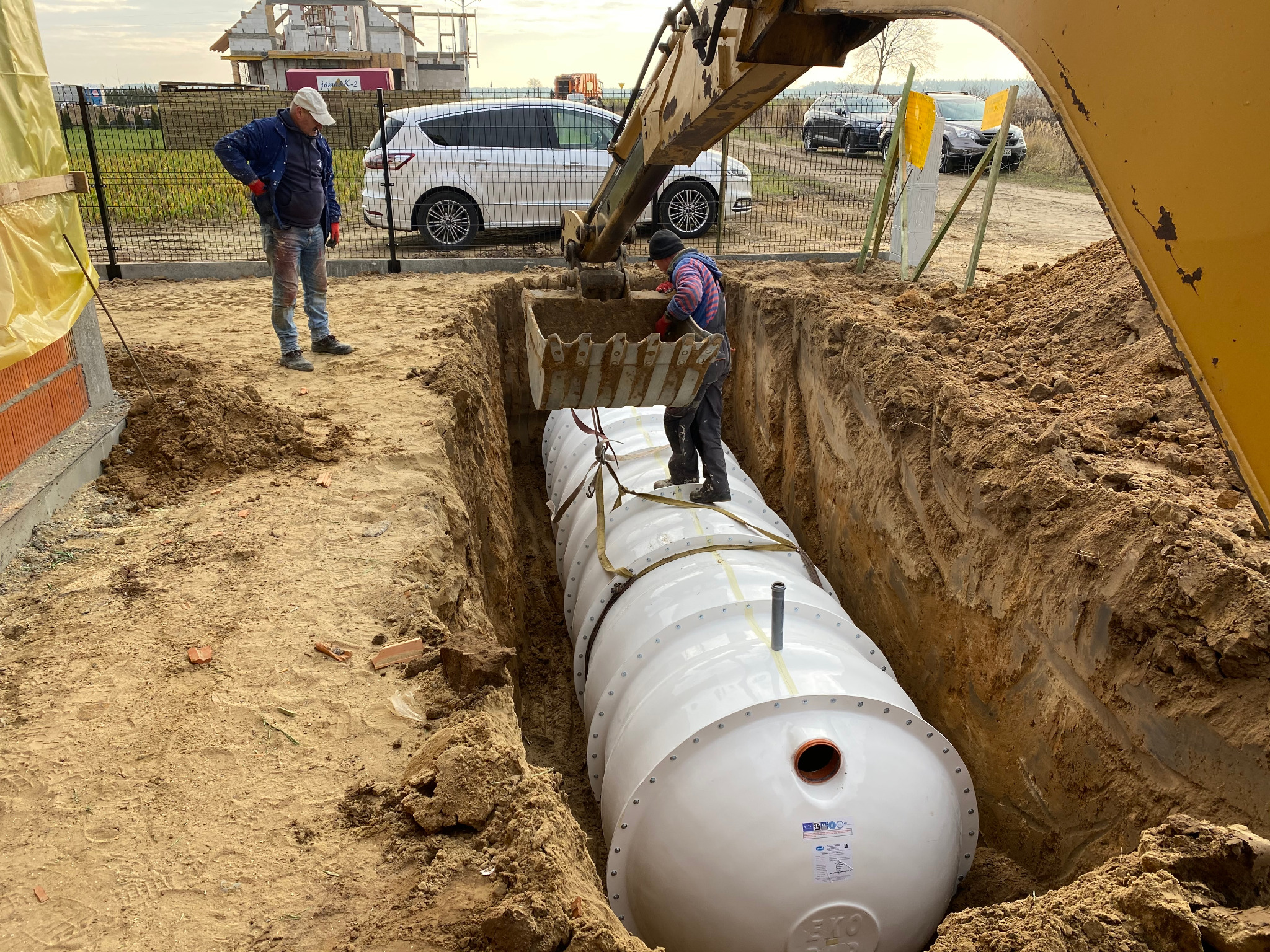 Instalacja białego, cylindrycznego zbiornika pod ziemią za pomocą koparki; pracownik w kasku i kombinezonie pomaga w osadzeniu zbiornika w wykopie, drugi pracownik obserwuje, w tle widoczny budynek...