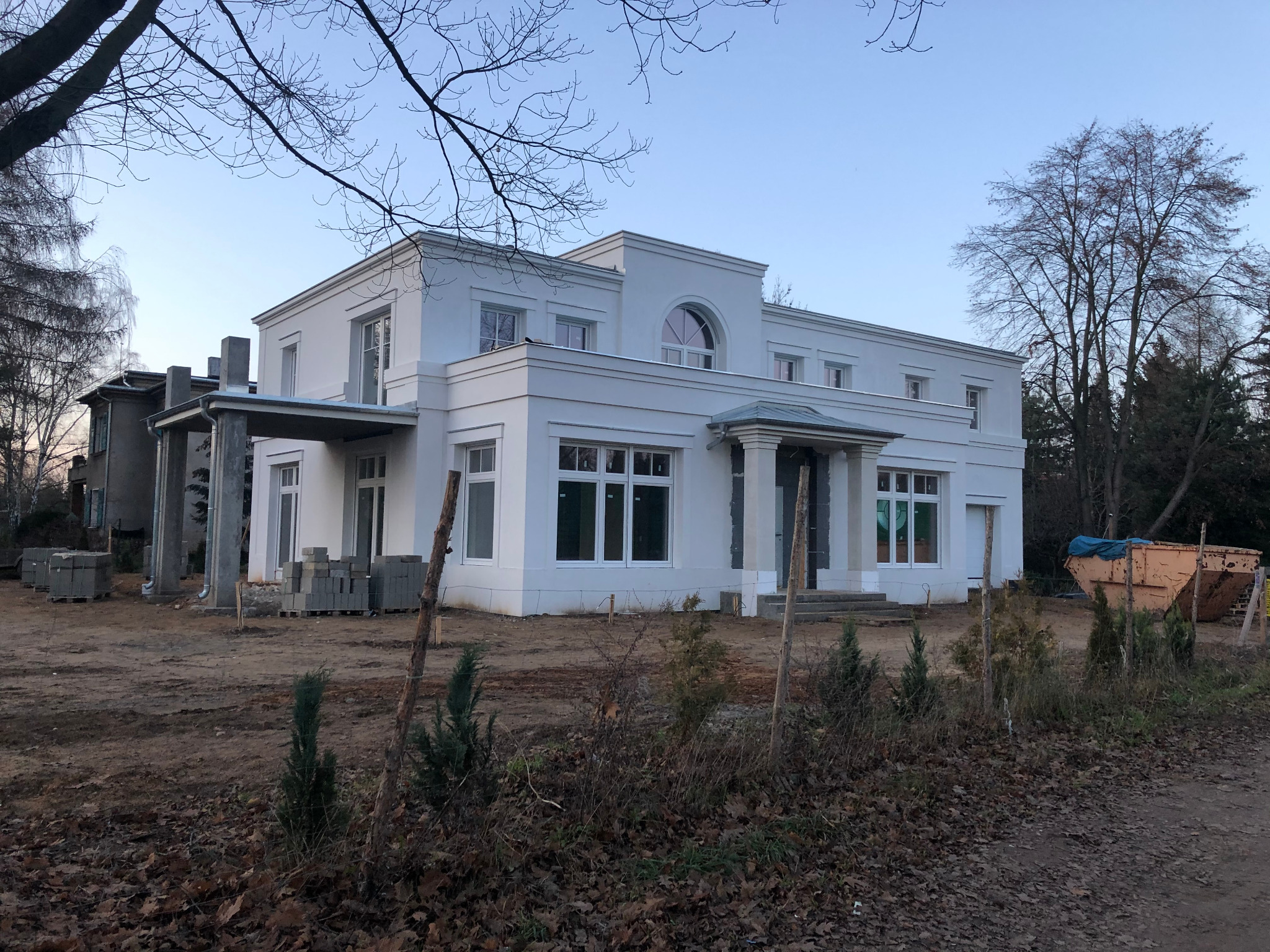 Biały, dwukondygnacyjny budynek z elementami klasycystycznymi w trakcie prac elewacyjnych, widoczne materiały budowlane i kontener na gruz.