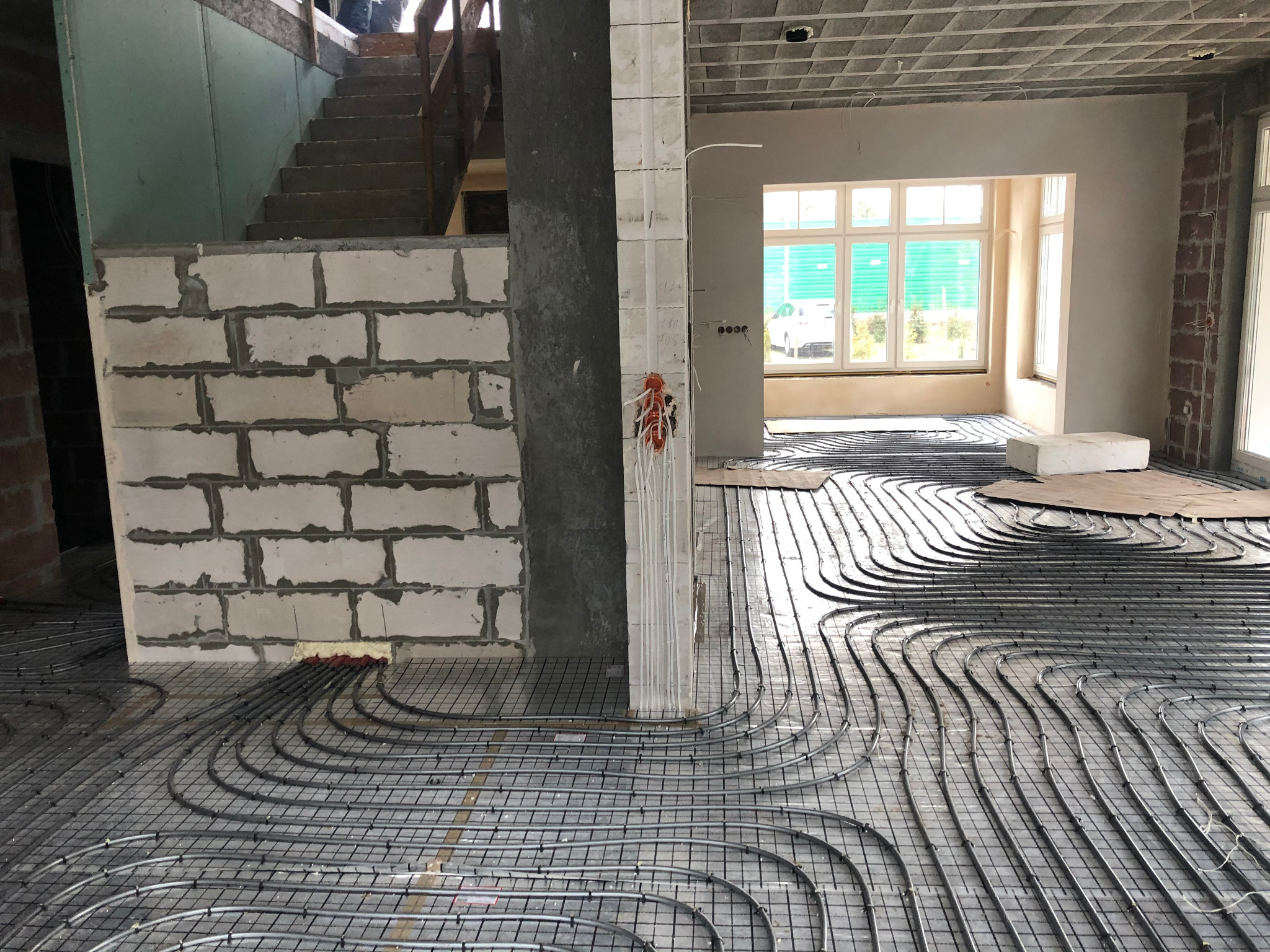Instalacja ogrzewania podłogowego w surowym wnętrzu, widoczne rury grzewcze ułożone na siatce, ściany z bloczków betonowych, instalacja elektryczna w puszce, sufit w stanie surowym.