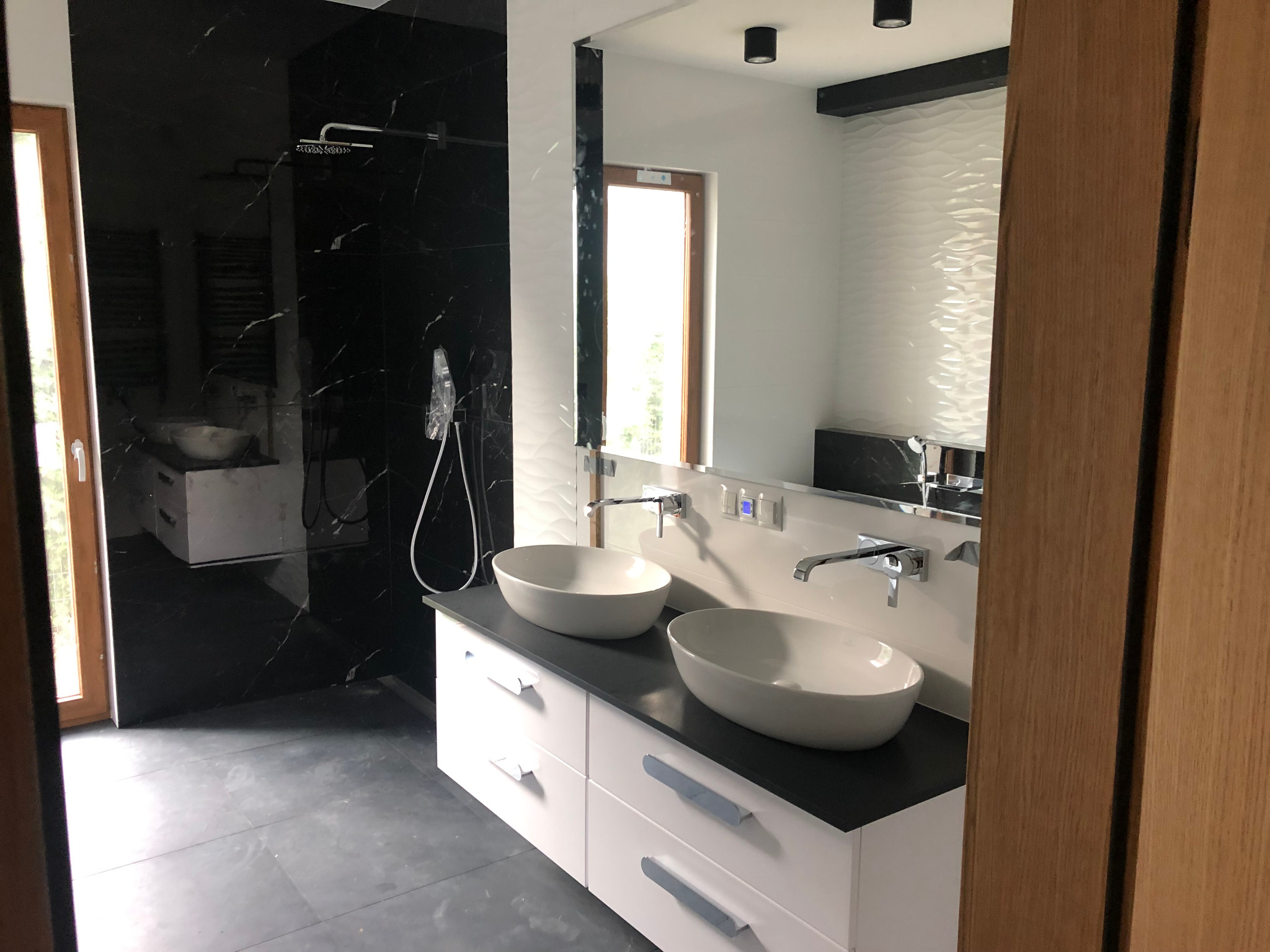 Nowoczesna łazienka z czarną marmurową ścianą prysznicową, dwiema owalnymi umywalkami nablatowymi na białej szafce z szarym blatem oraz lustrem odbijającym okno i białą ścianę z dekoracyjną fakturą.