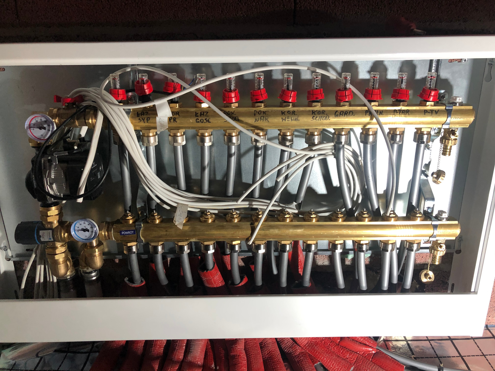 Rozdzielacz ogrzewania podłogowego w białej szafce, widoczne mosiężne rury z zaworami i manometrami, szare przewody oraz czerwona izolacja na rurach w dolnej części.