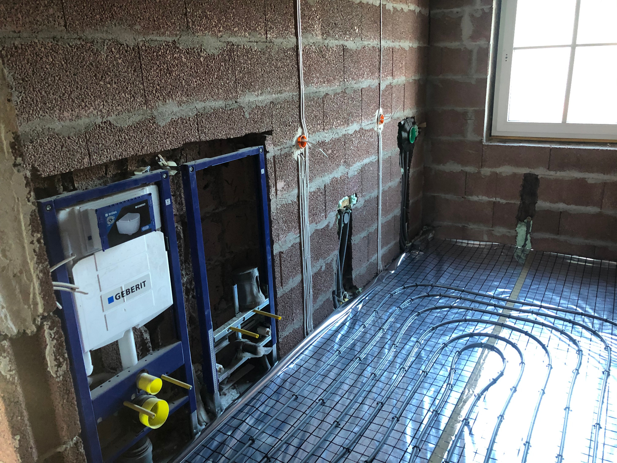 Instalacja ogrzewania podłogowego z rurkami na metalowej siatce, widoczne przewody elektryczne w ścianie z czerwonej cegły oraz zabudowa podtynkowego systemu sanitarnego Geberit z żółtymi elementami.