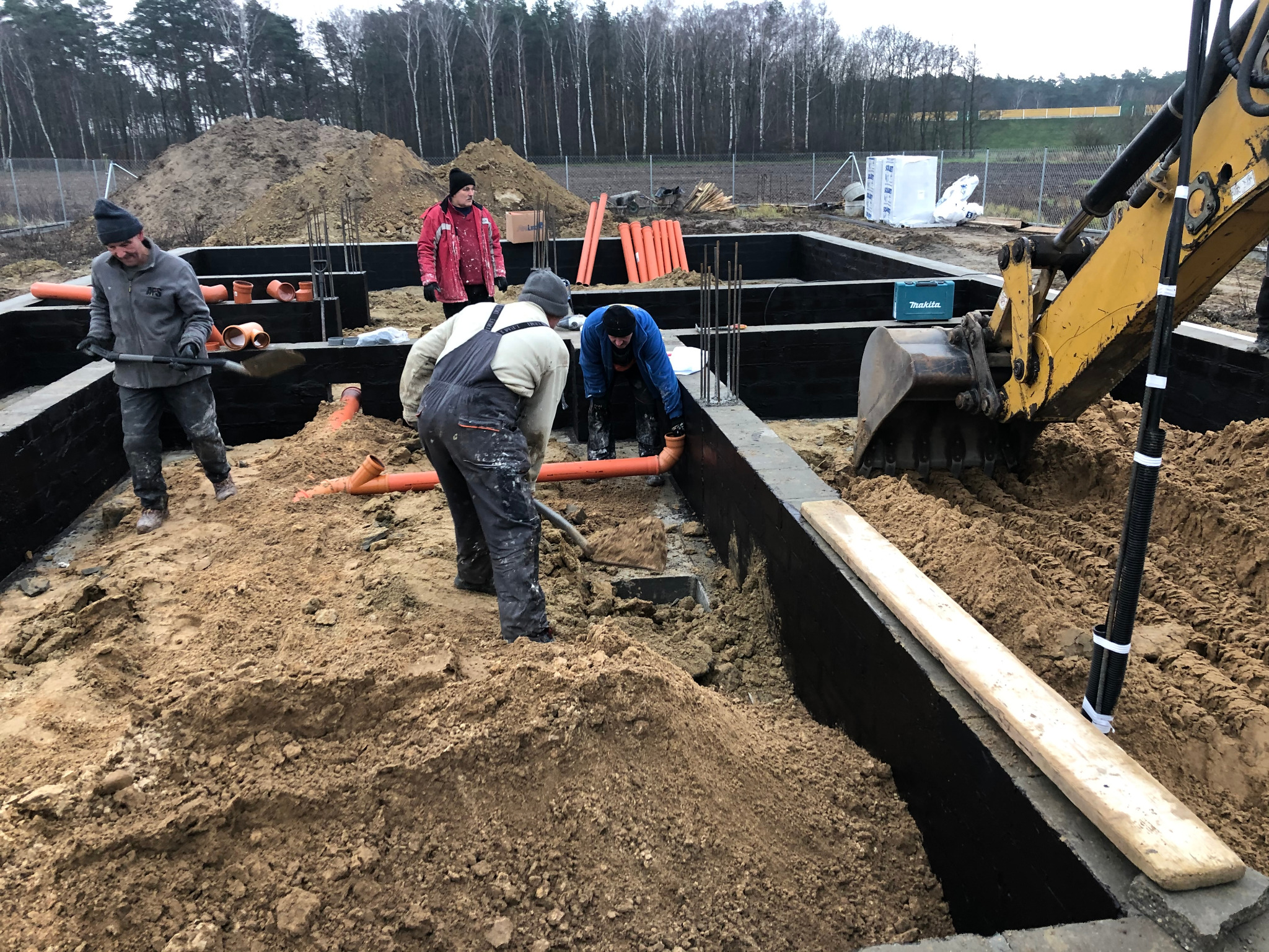 Prace ziemne przy fundamentach budynku: robotnicy układają rury kanalizacyjne w wykopie zabezpieczonym czarną izolacją, obok stoi żółta koparka i leżą sterty ziemi.