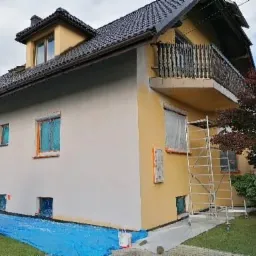 Dwukondygnacyjny dom z balkonem w trakcie odświeżania elewacji, część ściany pomalowana na biało, część na żółto, okna zabezpieczone folią, na trawniku rozłożona niebieska plandeka, przy ścianie...