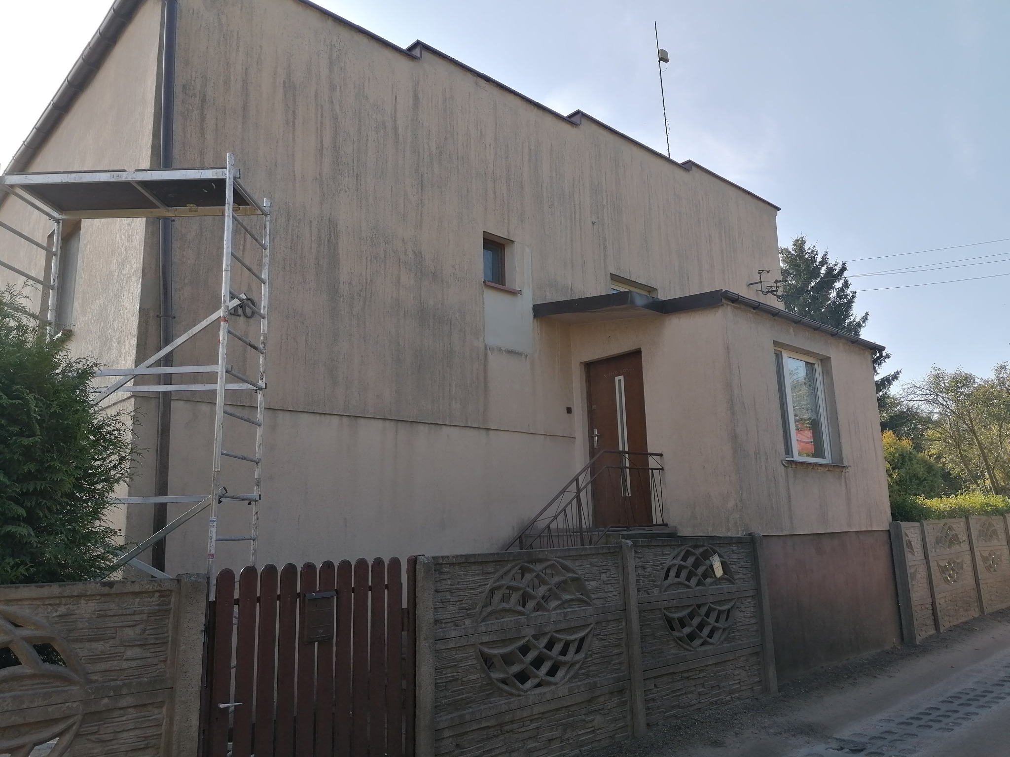 Dwupiętrowy dom z elewacją w odcieniach beżu, widoczne rusztowanie z lewej strony, betonowe ogrodzenie z dekoracyjnymi elementami, schody prowadzące do drzwi wejściowych.