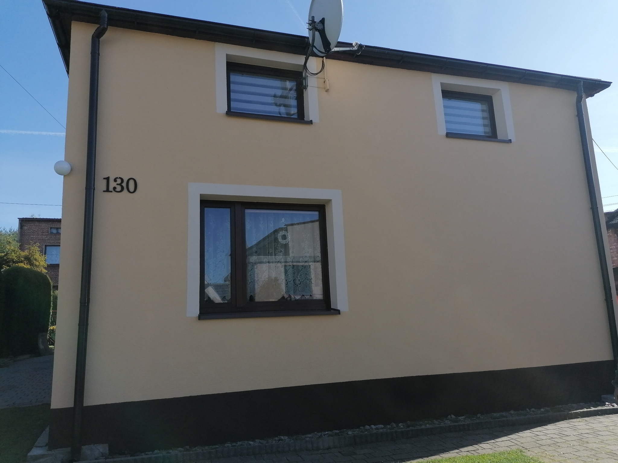 Dwukondygnacyjny budynek z beżową elewacją, ciemnym pasem u dołu, numerem domu 130, trzema oknami z białymi ramami i anteną satelitarną na górze, sfotografowany w słoneczny dzień.