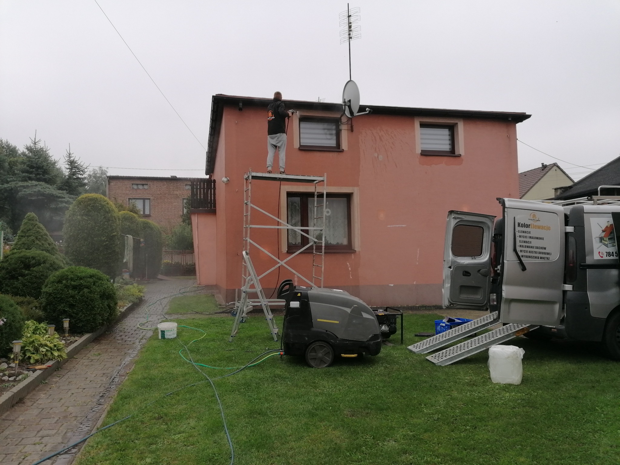 Pracownik na rusztowaniu czyści elewację domu za pomocą myjki ciśnieniowej, obok zaparkowany samochód dostawczy z logo firmy.
