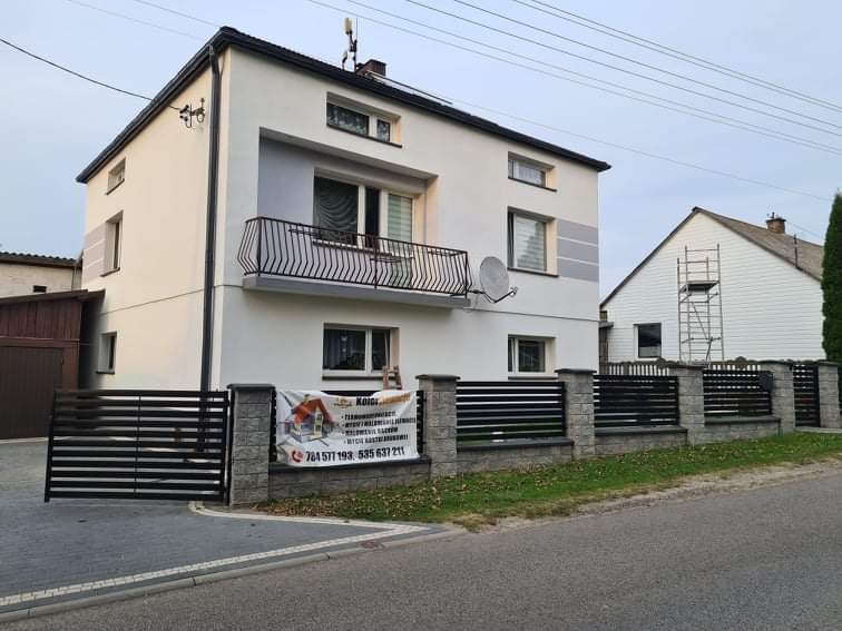 Dwupiętrowy dom z balkonem, pomalowany na biało z szarymi akcentami, widoczna brama i ogrodzenie, w tle dom z elewacją w trakcie prac z użyciem rusztowania.