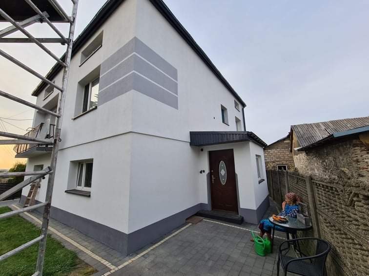 Dwupiętrowy dom po renowacji z białymi ścianami i szarymi akcentami dekoracyjnymi, widoczne rusztowanie, kobieta siedząca przy stoliku przed wejściem