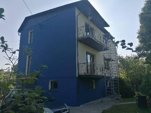 Dwukondygnacyjny budynek z balkonami po renowacji, z niebieską elewacją na części ściany i rusztowaniem przy balkonie, widok z poziomu gruntu, z samochodem na pierwszym planie.