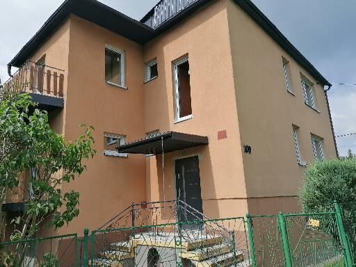 Dwupiętrowy dom z beżową elewacją, widoczne schody wejściowe z metalową barierką, okna z białymi ramami i zielone ogrodzenie.