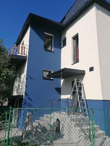 Dwukolorowa elewacja domu z drabiną opartą o ścianę, widok zza zielonego ogrodzenia.