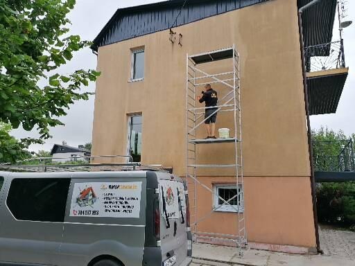 Pracownik na rusztowaniu maluje elewację dwukondygnacyjnego domu, widoczny fragment firmowego busa z reklamą.