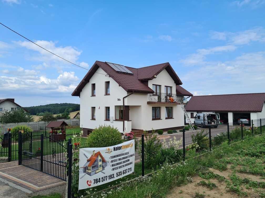 Dom jednorodzinny z brązowym dachem, po termomodernizacji, z widoczną drabiną przy balkonie, zaparkowanym busem i banerem reklamowym firmy zajmującej się elewacjami.