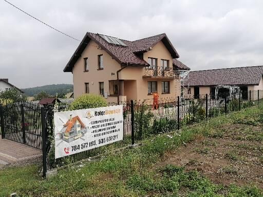 Dom jednorodzinny w Łazach po pracach remontowych, z widoczną elewacją w odcieniach beżu, ogrodzony czarnym metalowym płotem, na którym umieszczono baner reklamowy firmy.