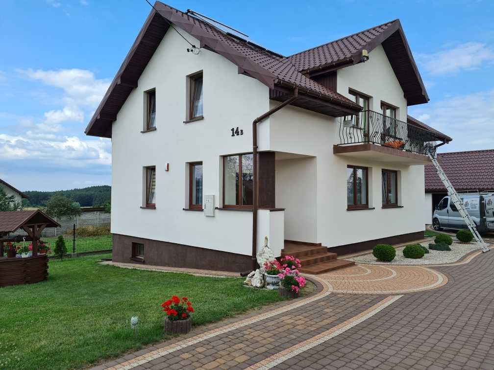 Biały dom z brązowymi akcentami, elewacja po renowacji, widoczny fragment podjazdu z kostki brukowej, drabina oparta o budynek, zadbany ogród z kwiatami.