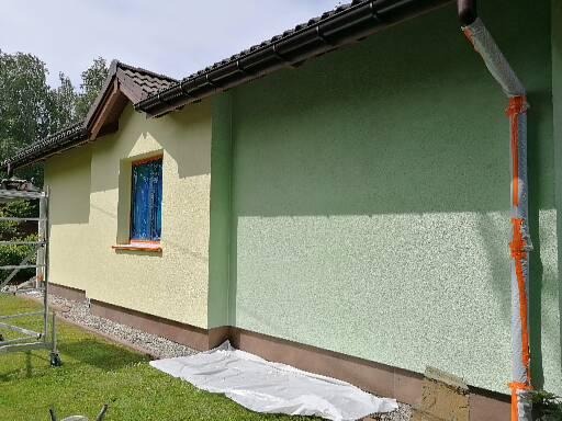 Dwu-kolorowa elewacja budynku z oknem zabezpieczonym folią, widoczny fragment rynny owiniętej folią ochronną i rozłożona plandeka na trawie.