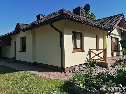 Jasny, parterowy dom z brązowym dachem i stolarką okienną, otoczony zielonym trawnikiem i kamiennym obrzeżem rabatki, widok z zewnątrz w słoneczny dzień.