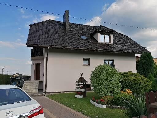 Dom jednorodzinny z nową, jasną elewacją, ciemną dachówką i zadbanym ogrodem z elementami dekoracyjnymi.