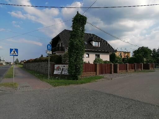 Dom jednorodzinny z ciemnym dachem i jasną elewacją, otoczony drewnianym ogrodzeniem, z przodu widoczny chodnik i ulica, na ogrodzeniu wisi baner reklamowy.