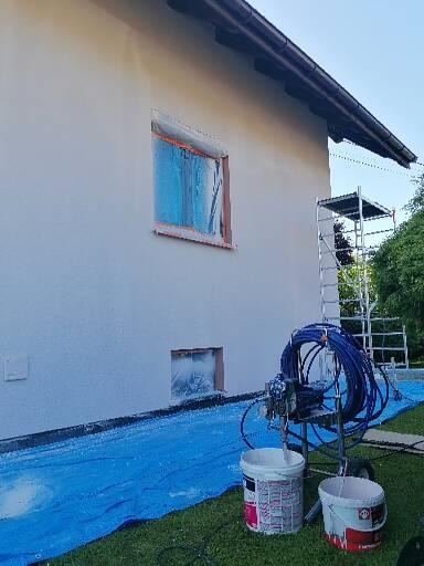 Elewacja domu w trakcie natryskowego nanoszenia farby, okna zabezpieczone folią, widoczny agregat malarski, dwa wiadra z farbą i rozłożona folia ochronna.