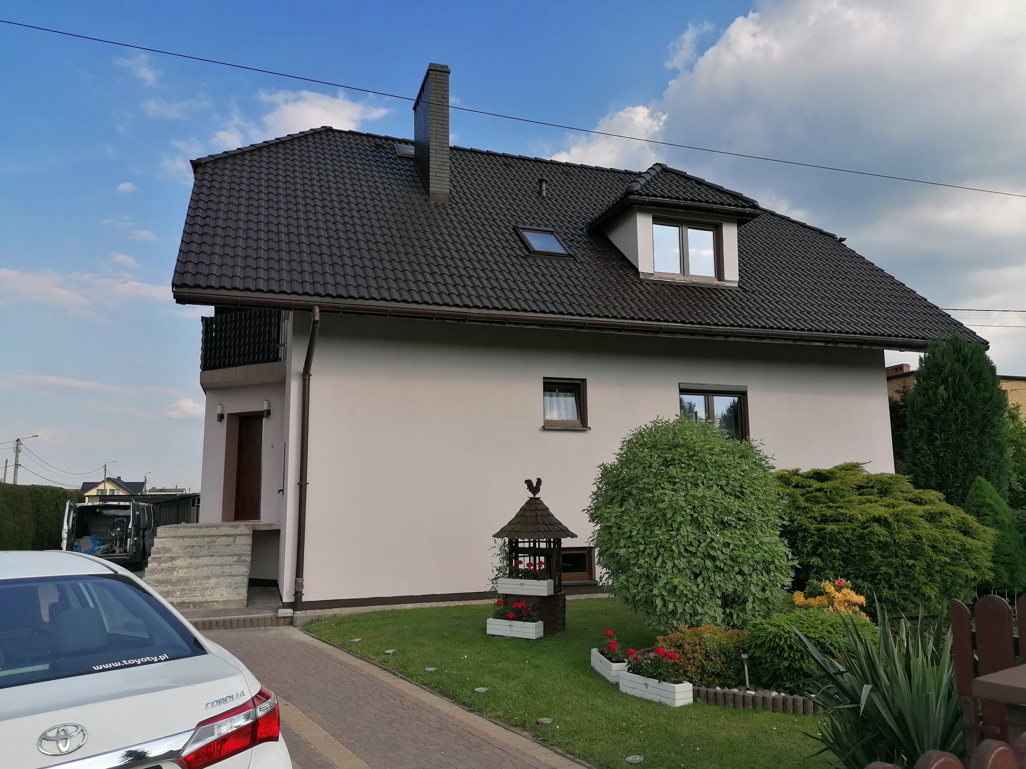 Dom jednorodzinny z ciemną dachówką, jasną elewacją i zadbanym ogrodem, widoczny z perspektywy chodnika z kostki brukowej, z samochodem osobowym zaparkowanym na podjeździe.