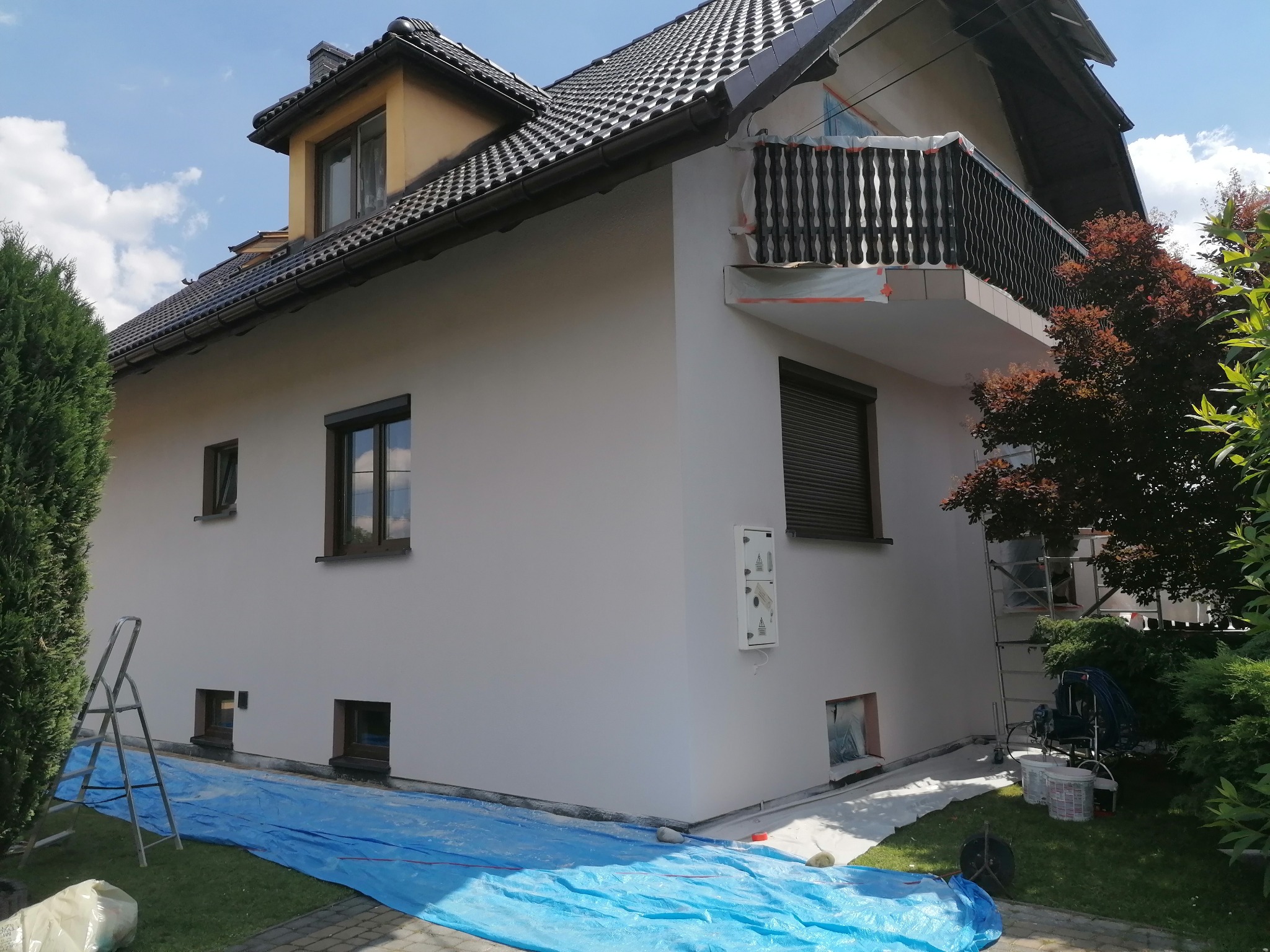 Malowanie elewacji domu jednorodzinnego w trakcie realizacji, z zabezpieczonymi oknami i podłożem folią, widoczny sprzęt do malowania natryskowego i drabina.