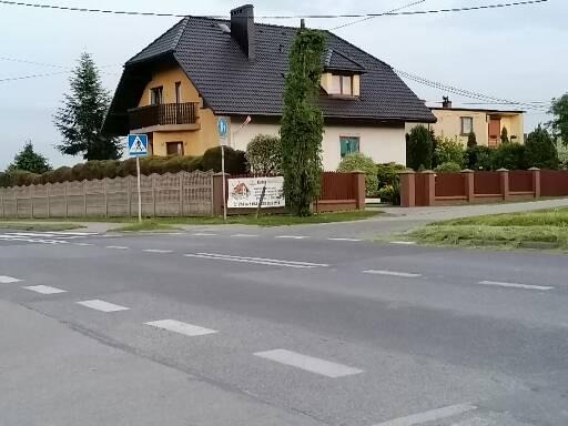 Żółty dom jednorodzinny z ciemnym dachem widoczny z ulicy, ogrodzony betonowym i drewnianym płotem, z reklamą firmy budowlanej na ogrodzeniu.