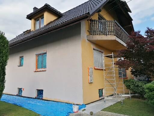 Dwukondygnacyjny dom z balkonem w trakcie odświeżania elewacji, część ściany pomalowana na biało, część na żółto, okna zabezpieczone folią, na trawniku rozłożona niebieska plandeka, przy ścianie...