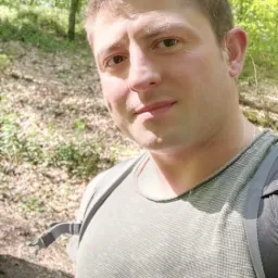 Portret mężczyzny z plecakiem na tle leśnym, z widocznym uśmiechem i zmarszczkami mimicznymi.