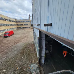 Montaż stalowej belki nad otworem w hali przemysłowej, widoczne rusztowania wewnątrz, na zewnątrz czerwony bus i budynek w tle.