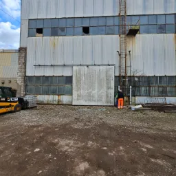 Pracownik w pomarańczowych spodniach przy metalowej bramie starej hali przemysłowej z widocznym wózkiem widłowym JCB i stertą metalowych elementów.