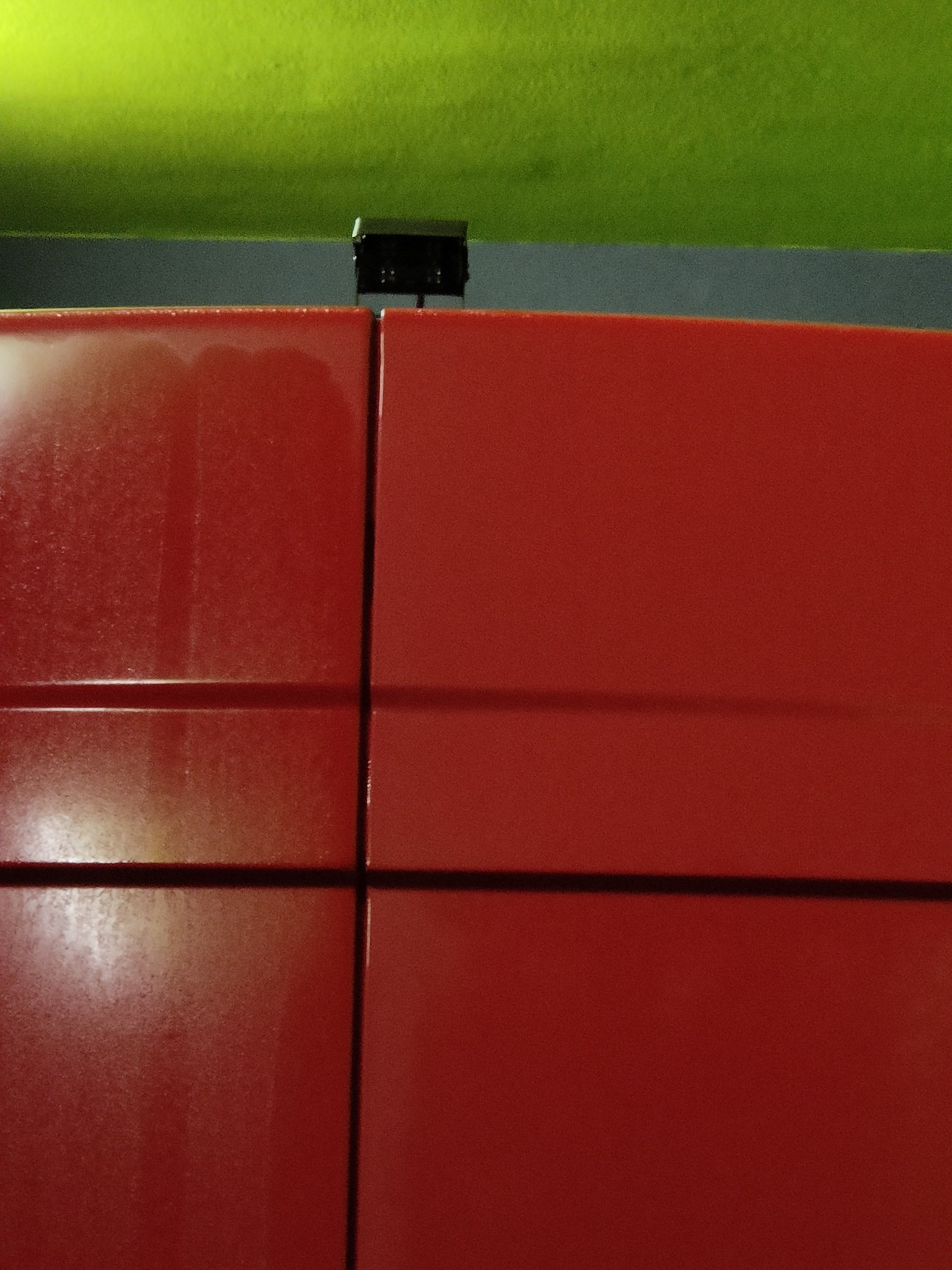 Fragment czerwonej, metalowej konstrukcji z widocznymi spawami, na tle szarej i zielonej ściany, oświetlony górnym światłem.