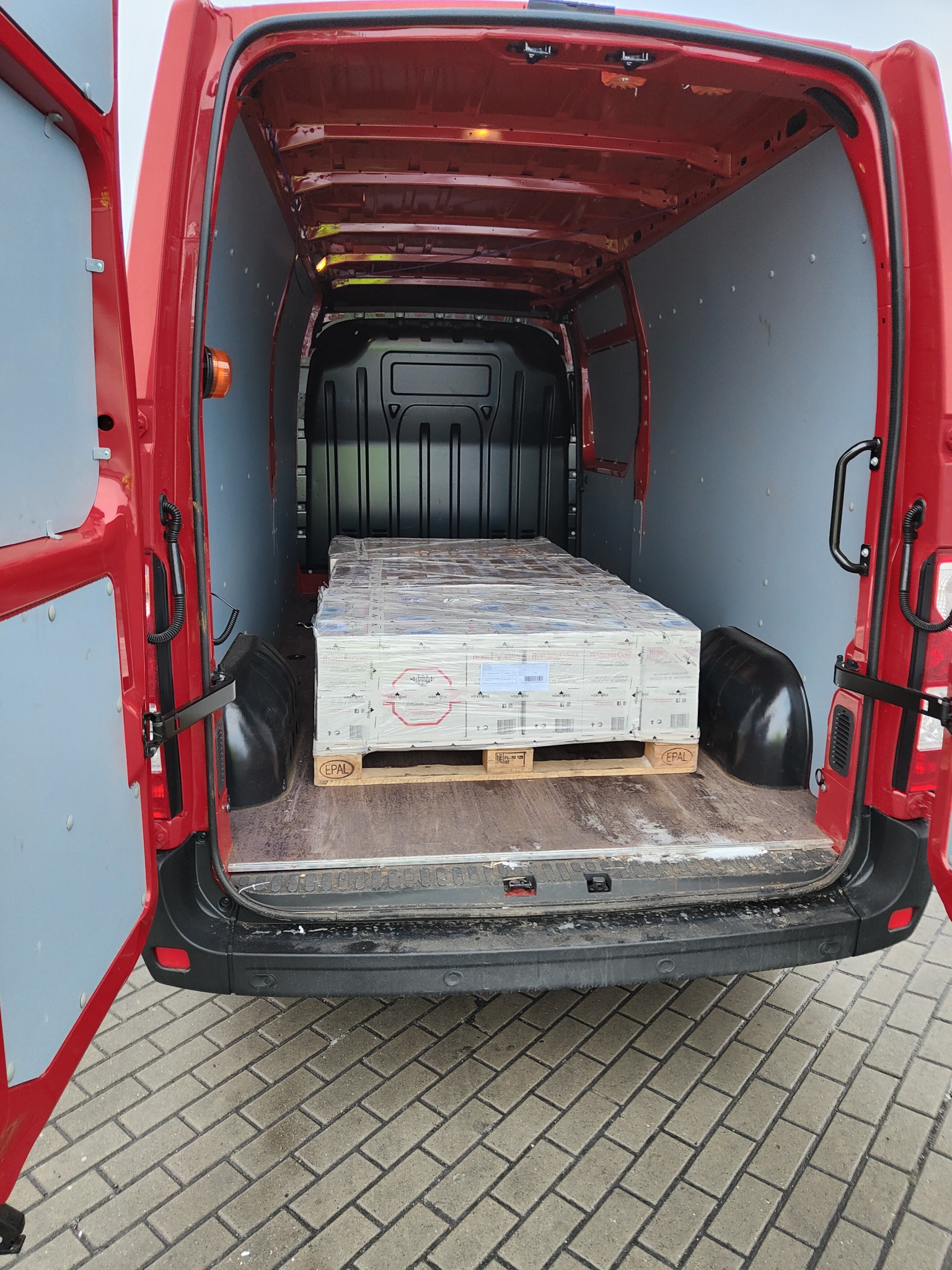 Wnętrze czerwonego busa z otwartymi drzwiami, załadowane paletą z zapakowanymi kartonami, widok od tyłu.