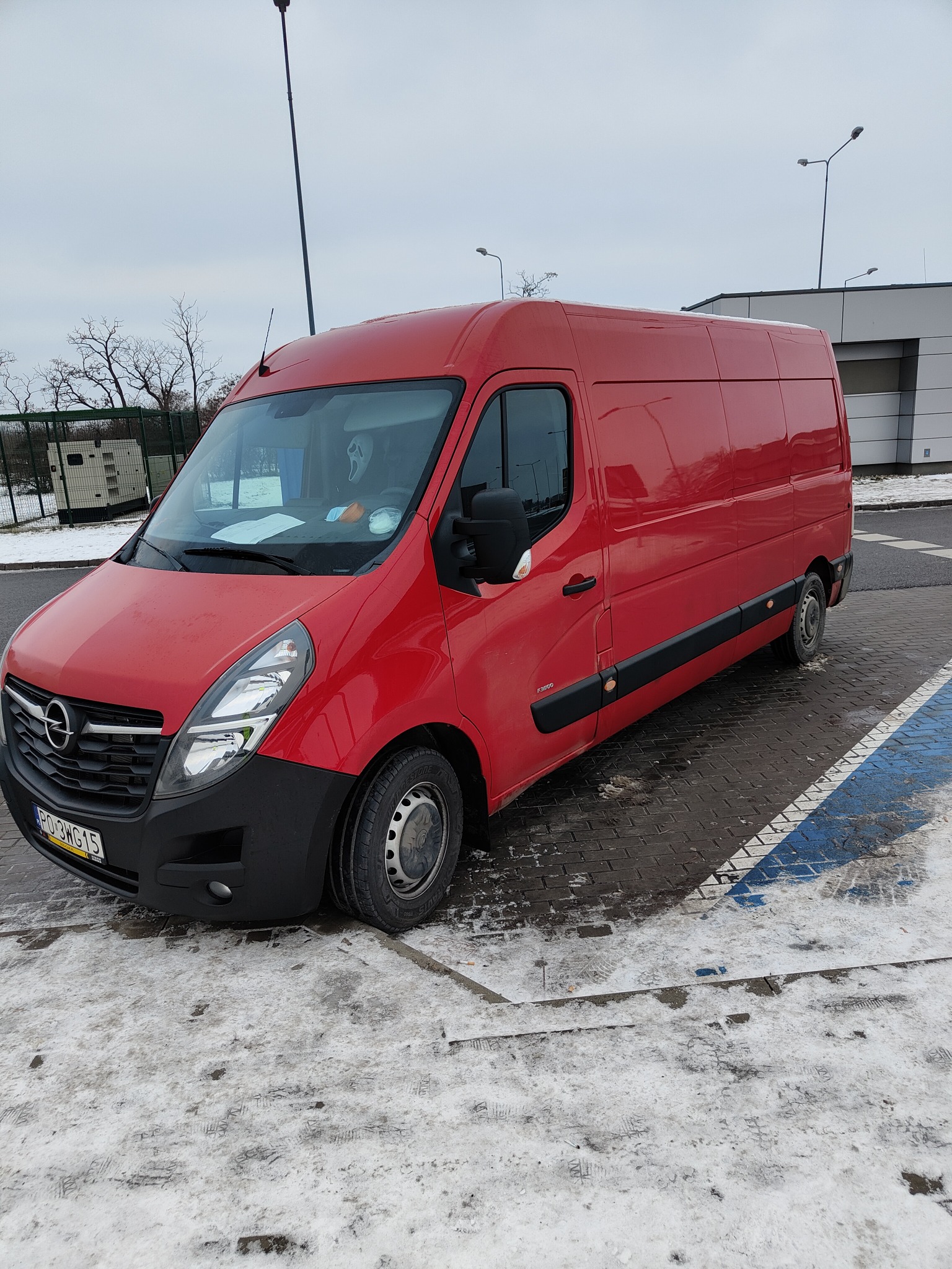 Czerwony samochód dostawczy Opel Master zaparkowany na parkingu w zimowych warunkach, widoczne ślady śniegu i błota na kostce brukowej, w tle budynek z szarymi panelami.
